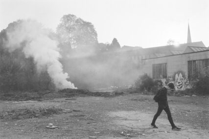 Black and white view of a youth striding off to the right of an open area while to the left in the background a bonfire burns, a thick column of smoke rising into the late afternoon light.