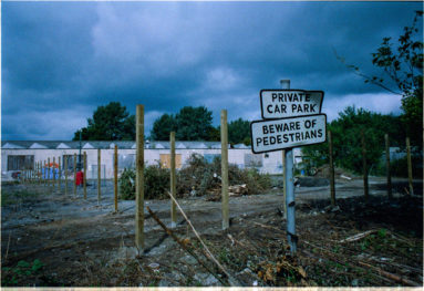 In the foreground a battered sign reads "Private care park. Beware of Pedestrians". Bare wooden upright posts lead away to the background to low, old industrial sheds and a brooding sky.