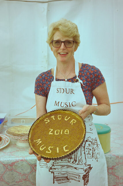 A bespectacled woman in a Stour Music Festival apron holds a tart for the view of the camera with the words Stour Music 2018 written out in pastry on its surface.