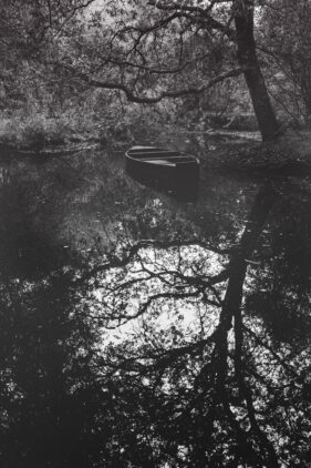 A small wooden boat sits on a tree-lined section of water, the trees reflected in the water.