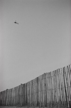 A wooden stick fence dominates the foreground as a helicopter, very small in the frame, crosses the clear sky above.