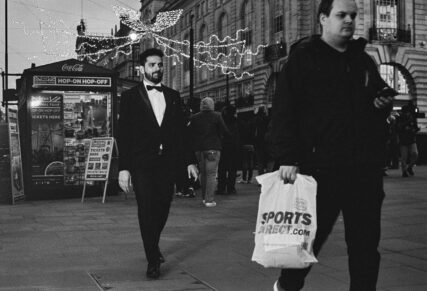 A man in the foreground walking to the right of the frame, carrying a Sports Direct plastic bag, is followed by a smart bearded man in a tuxedo and bow tie.