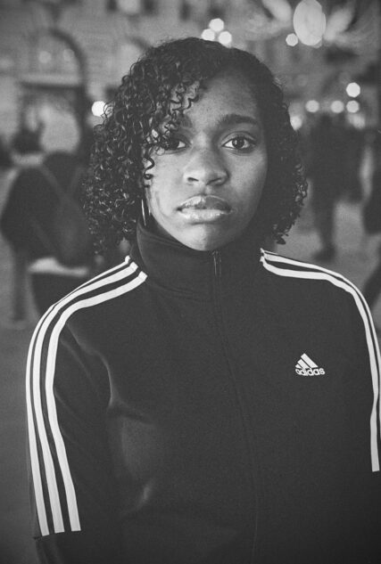 Black and white portrait of a black girl with curly hair looking directly at the camera wearing an Adidas sports top.