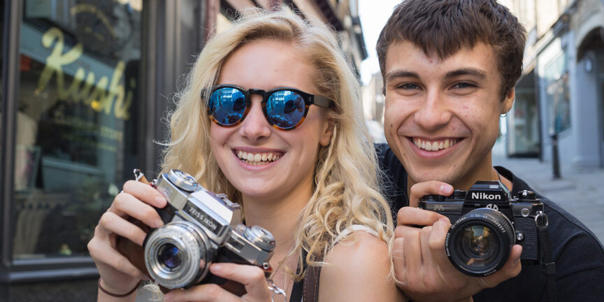 A young blonde female and her brunette boyfriend holding cameras and smiling at the camera.