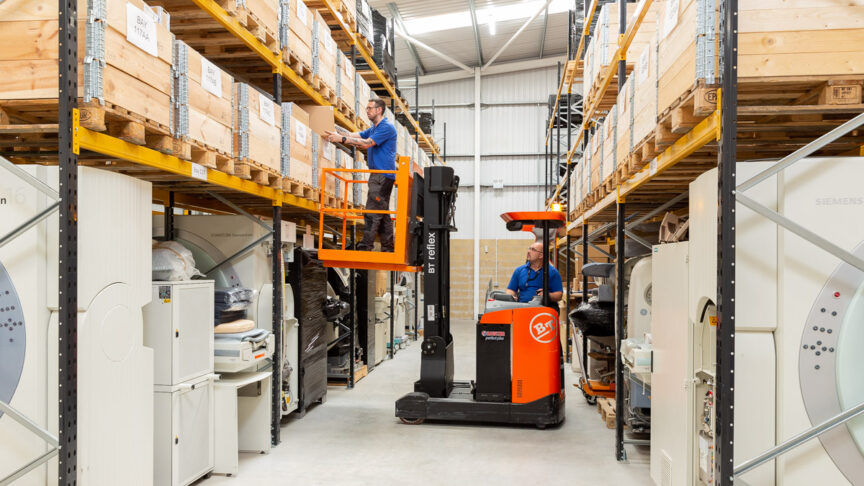 A warehouse man stands in an elevated platform to reach a box on a high shelf while the lift operator looks on from the cabin of the elevator truck.