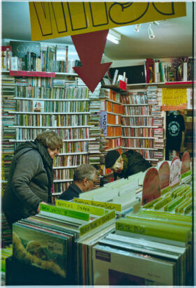 A man and two women look down behind a rack of vinyl records to search in more boxes of records for a birthday gift.