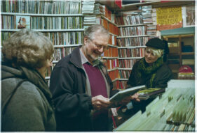 A man smiles, looking down at a vinyl record while his wife and possibly daughter look on.