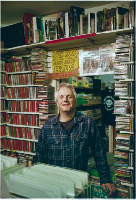 Portrait of Richard Churchyard, owner of Raves from the Grave record shop in Frome, with records and CDs visible all around him.