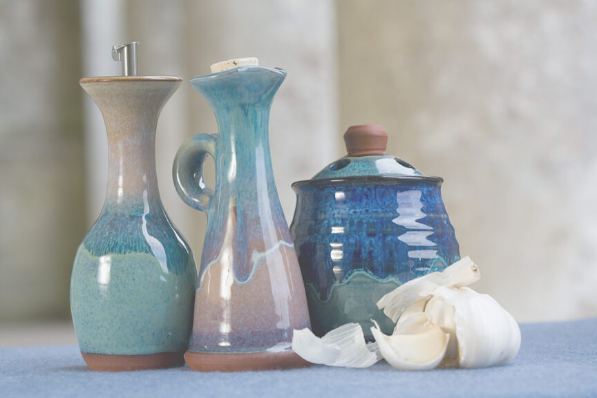 Three ceramic products in a row, an oil bottle, a jug and a garlic roaster, on a blue cloth with broken garlic bulbs lying next to them.