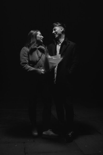 black and white photo of a young male and female couple standing looking at each other. She is holding flowers wrapped in paper.