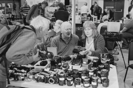 A man and a woman running one of the camera stalls help a male customer looking at a Polaroid camera.