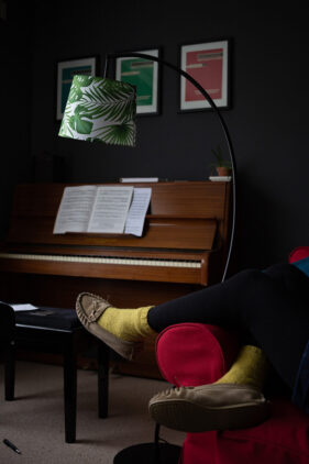 A piano in the background with someone's feet in the foreground, wearing yellow socks and resting on the red arm of a comfy chair. I green leaf-pattern lamp hangs above.