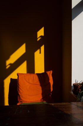 Window light casts the shadow of the window frame onto a bright yellow wall. A chair in front of the wall sports a bright, burnt orange cushion.