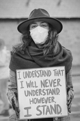 A stylish young woman in a dark, wide-brimmed hat and white face mask hold a banner which reads, "I understand that I will never understand. But I stand"