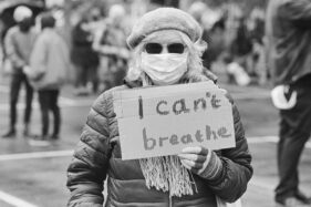 An older female protestor carries a piece of cardboard on which is written, "I can't breathe"