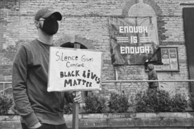 A male protester wearing a black mask carries a placard which reads "Silence gives consent. Black lives matter". A banner on the building behind him reads "Enough is enough."