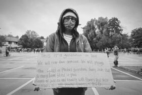 A young protester carries a large cardboard sign which has an extended hand-written protest text on it.