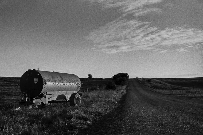 Black and white photo of a farm tanker parked on the edge of a field with a mettled track leading past and to the horizon on a sunny evening.