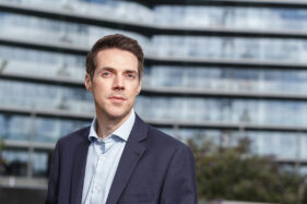 Portrait of a male business man looking to camera with the strong horizontal lines of an office block blurred behind him.