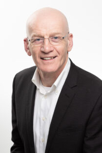 A bald gentleman wearing glasses, dark jacket and open-necked shirt smiles gently to camera.