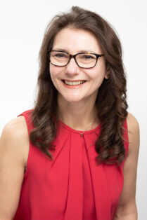 A professional female wearing glasses, wearing a red top and smiling off-camera.