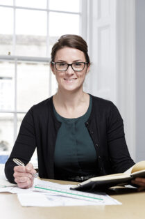 A professional woman sits at her office desk, pen in hand, looking to camera and smiling.