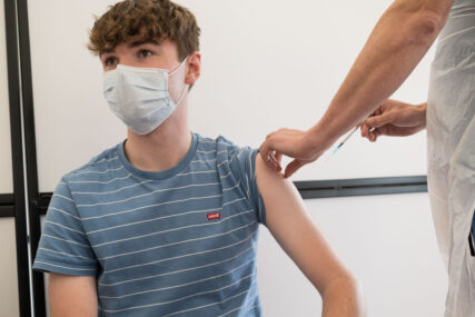 A young man receives his Covid vaccination. He is wearing a mask and we can see the nurse's hands, one of which has just withdrawn the syringe.