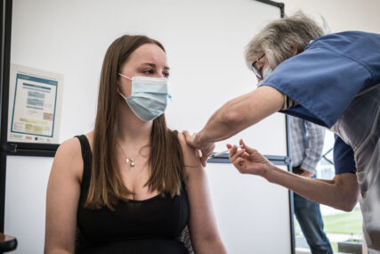 A young woman receives her Covid vaccination. She is wearing a mask and we can see the nurse holding the upper arm into which she has inserted the syringe.