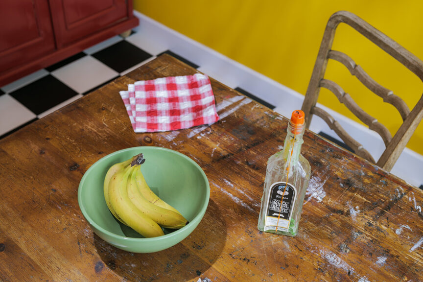 A still life looking down on a paint-smeared wooden table with a green bowl of yellow bananas with a red-chequered napkin behind and a candle in a bottle to the right of the bowl. A black and white chequered floor, yellow wall and bottom of a red cupboard are visible beyond the edges of the table. beyond