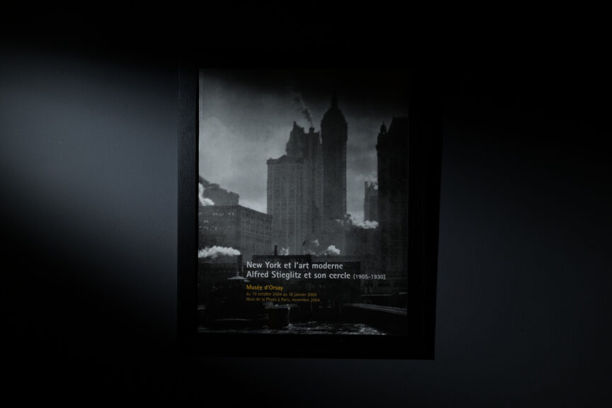 An Alfred Stieglitz poster framed in black on a grey wall is strongly lit from one side.