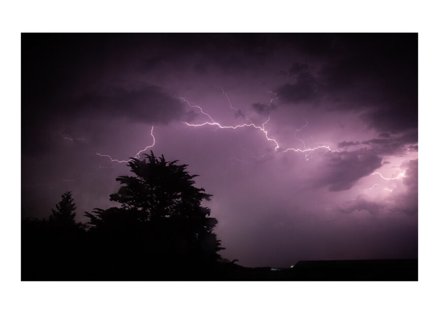 Lightning at night silhouettes a large conifer tree and low factory roofs against a cloudy, purple sky.