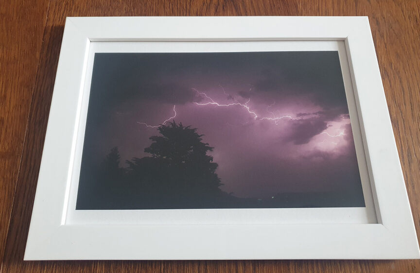 A print framed in white of a dark, purple night sky illuminated by lightning with a large confer tree and low factory roofs silhouetted.