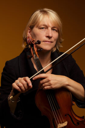 Colour photo of Viola player Rachel Byrt wearing a black coat holding her viola and looking to camera.