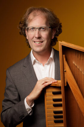 Colour portrait of harpsichord player Silas Wollston holding his virginal and looking to camera.