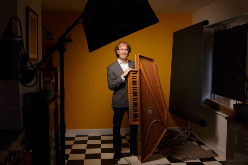 Photo showing Silas Wollston standing with his up-ended virginal, with the photographer's studio light and "flag" in shot.