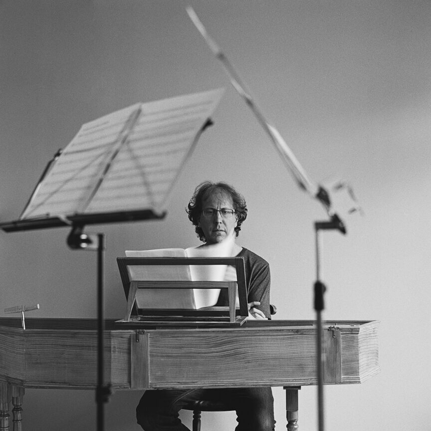 Black and white square format photo of Silas Wollston siting at his virginal (harpsichord), seen through two music stands which are in the foreground.