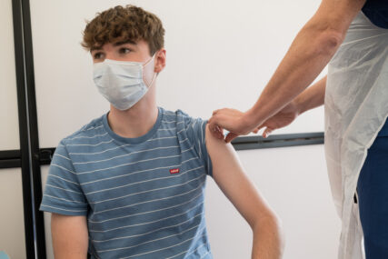 A young man is seen receiving his vaccine. We can only see the man, who is wearing a mask, and the arms of the nurse administering the vaccine.