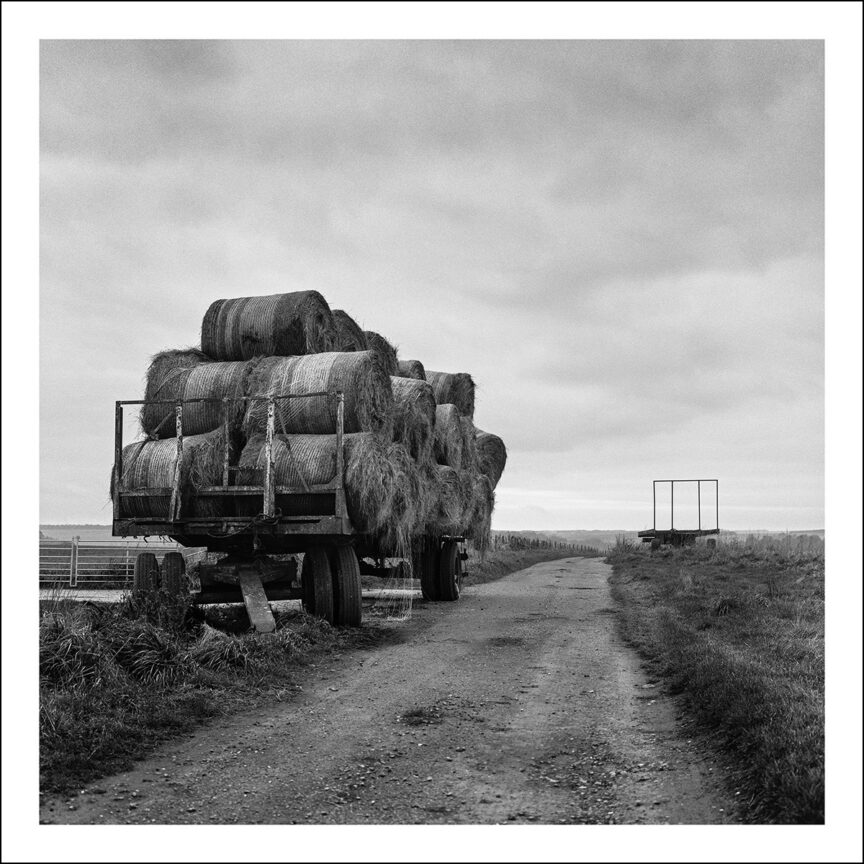 A waggon is laden with round hay bales with a farm track running past.