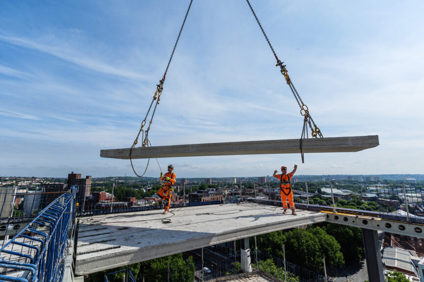 Two construction workers stand on a concrete floor as another section of floor is craned into position. They reach up to guide it into place with a view of the Bristol skyline visible behind them on a sunny day.