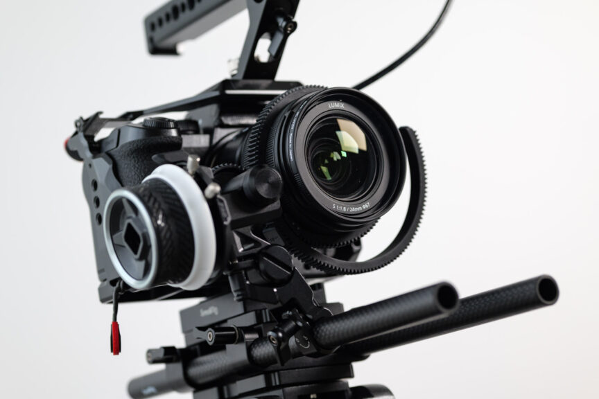 Angled close-up photo of a mirrorless camera mounted on a video tripod with a follow-focus device attached.