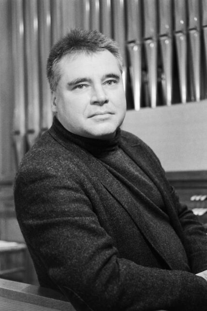 Black and white portrait of musician Ralph Stelzenmüller looking to camera with organ pipes in the background.