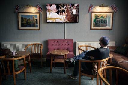 A large wall-mounted television is showing the moment King Charles III is crowned. There is just one man sitting watching the event in a side bar where all the other chairs are empty.