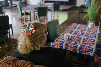 View of the back of a woman's head. She has curly strawberry blonde hair with a union jack hair clip in it with red, white and blue ribbons. She is seated at a table covered in a union jack oilcloth.