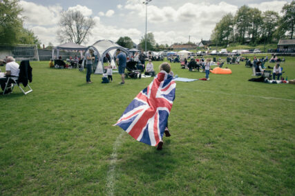 A young girl wearing a large union jack like a cape runs away from the camera across a field towards coronation picnickers.