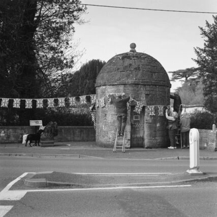 Black and white photo of people draping union jack bunting along a street and round a historical stone blind house (or jail) in Shrewton, Wiltshire. A black dog is standing watching what's going on.