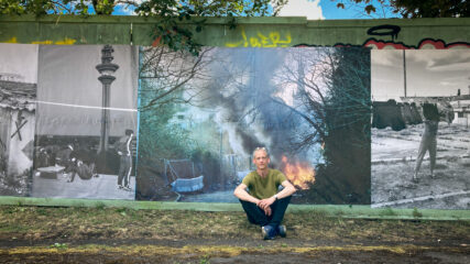 Photographer Tim Gander sits on the pavement in front of his print which is stapled to the hoardings. The photo behind him depicts the flames and smoke of a burning caravan.