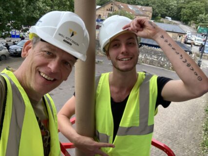 Selfie of me on the left and my son Joe taken on the scissor lift at full height as we work to uninstall the vinyl prints. Joe is doffing the peak of his hard hat and holding a cardboard tube we used to roll up the banner images.