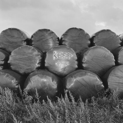 A stack of round, plastic-wrapped hay bales sitting on scrub grass with sky above and the words Keep Off spray-painted on the a bale in the centre of the photo.