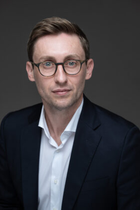 Head and shoulders portrait of a man in a dark blazer, open neck white shirt and glasses looks directly into the camera without smiling.