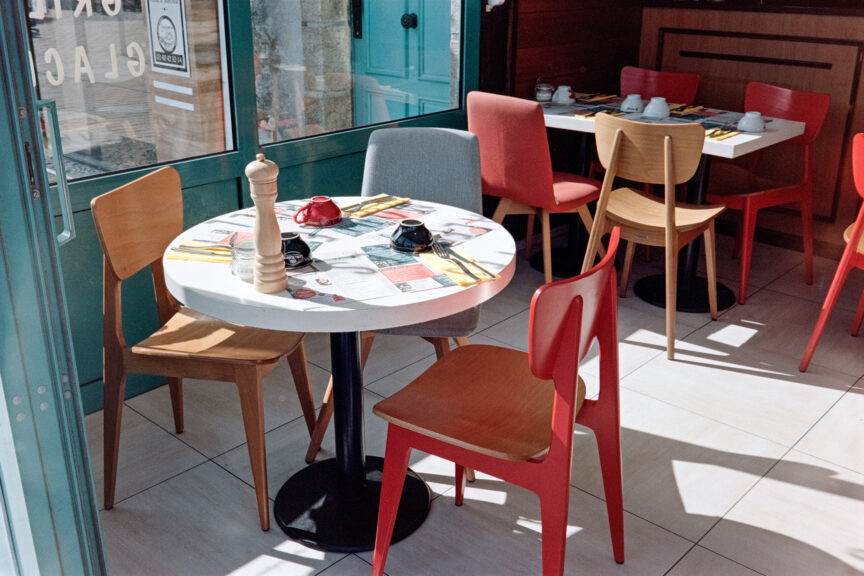 View into a French seaside cafe showing sunlight hitting a white table surrounded by red, grey and natural wood chairs. The table is set for three, with mats and coffee cups.